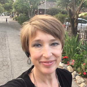 Head shot of me walking along a brick and concrete path lined with trees and flower gardens. I have a light brown pixie cut and dangly teardrop earings.